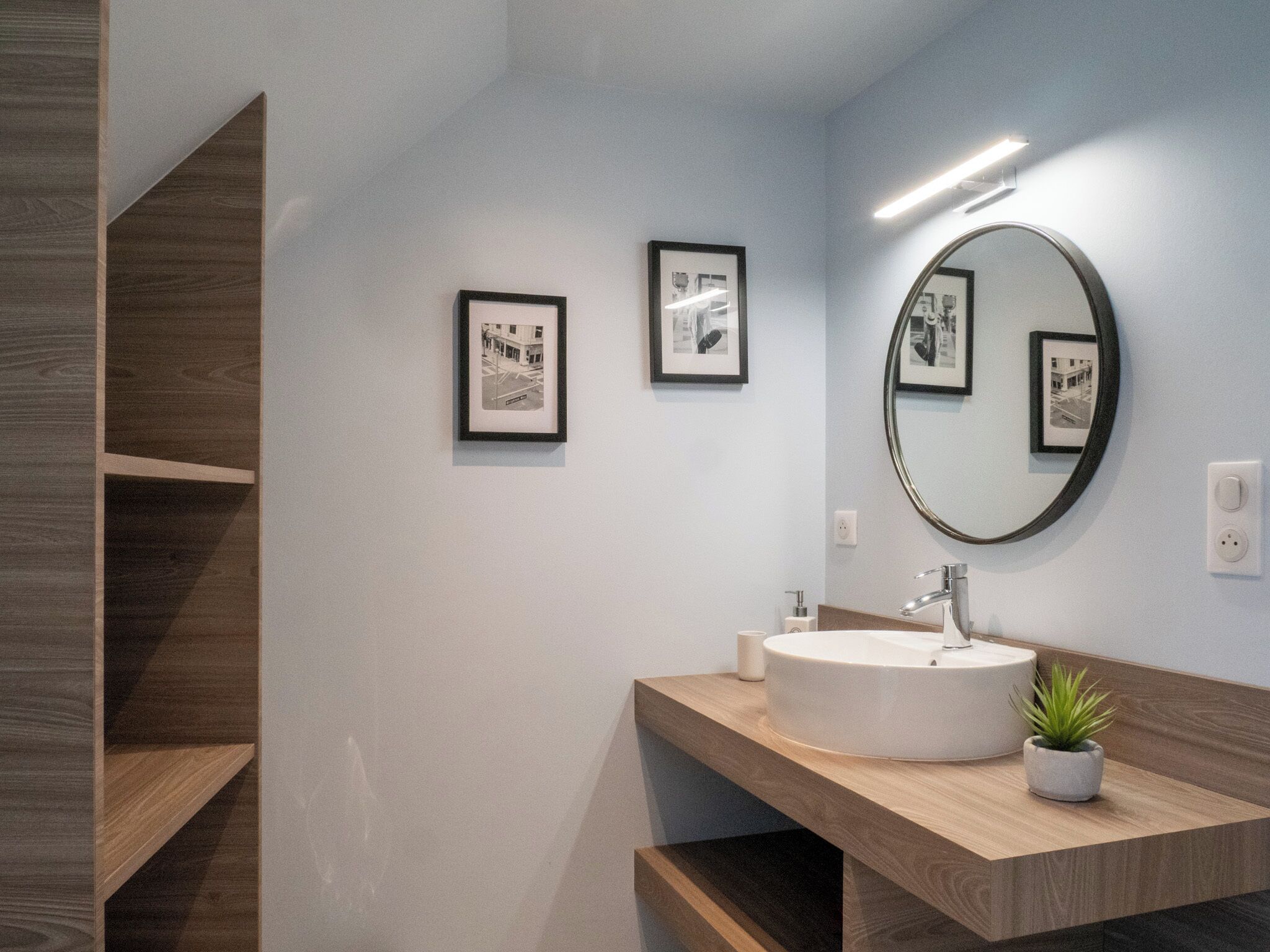 A modern bathroom with a round mirror, framed black and white photographs, and a wooden vanity.