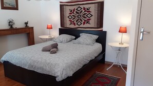 Ein ordentlich bezogenes Bett mit einem gemusterten Überwurf, zwei Nachttischlampen, einem Wandbild und einem kleinen Holztisch mit einer Lampe.