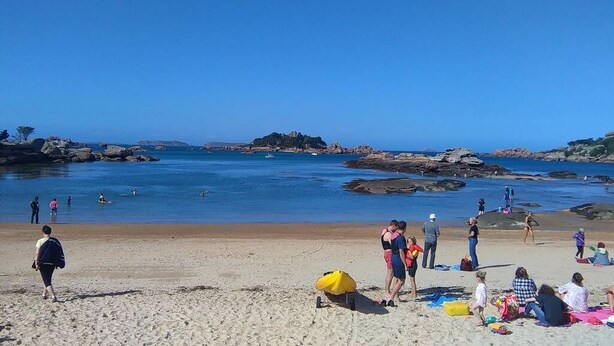 Uma praia com pessoas se banhando e tomando sol, céu azul límpido e afloramentos rochosos ao fundo.