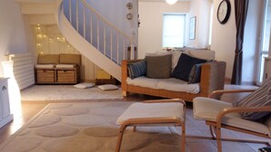 Uma sala de estar aconchegante com um sofá de vime, duas poltronas, um tapete branco, uma escada e uma pequena mesa com abajur.