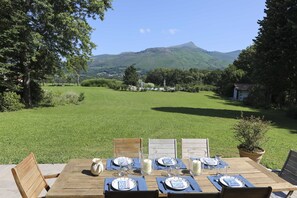 A wooden table set for four with plates, glasses, and a pitcher, overlooking a grassy field and a mountain.