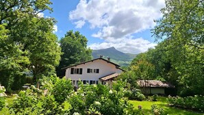 Een wit huis met een schuin dak, omringd door weelderige begroeiing en een berg op de achtergrond.