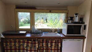 A compact kitchen with a microwave, coffee maker, and a small refrigerator. There are two windows providing natural light and a view of greenery outside. - Small charming Breton house (Nivillac)