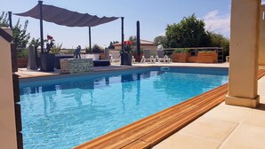Una piscina rectangular con borde de madera, rodeada de sillones de descanso y un muro de piedra.