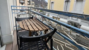 A balcony with a wooden table and chairs, overlooking a street with buildings and a mountain in the background.