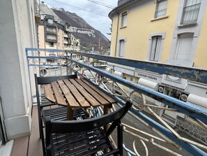 A balcony with a wooden table and chairs, overlooking a street with buildings and a mountain in the background. - Pied du télécabine. T2 + alcove. Parking couvert. (Cauterets)