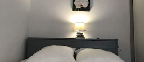 Un lit avec une literie blanche, une table de chevet avec une lampe et deux éléments de décoration murale.