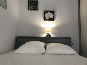 Una cama bien hecha con almohadas blancas y una sola almohada blanca en la cabecera. Una mesita de noche con una lámpara está colocada al lado de la cama.