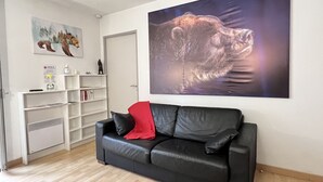 A living room with a black leather sofa, a red pillow, a white bookshelf, and a large bear painting on the wall.
