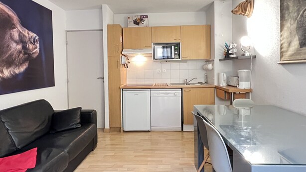 A compact kitchen with a microwave, dishwasher, and wooden cabinets. A black leather sofa with a red pillow is in the living area, and a glass table with chairs is near the kitchen.