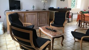 Una sala de estar con un mueble de madera, dos sillones negros, una mesita con un tablero de ajedrez, un televisor y un cuadro en la pared.
