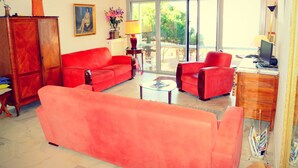 Ein Wohnzimmer mit einem roten Sofa, einem kleineren roten Sessel und einem Couchtisch. An der Wand hängt ein Gemälde, eine Lampe steht dort und ein Fenster bietet Blick auf Grünfläche draußen.