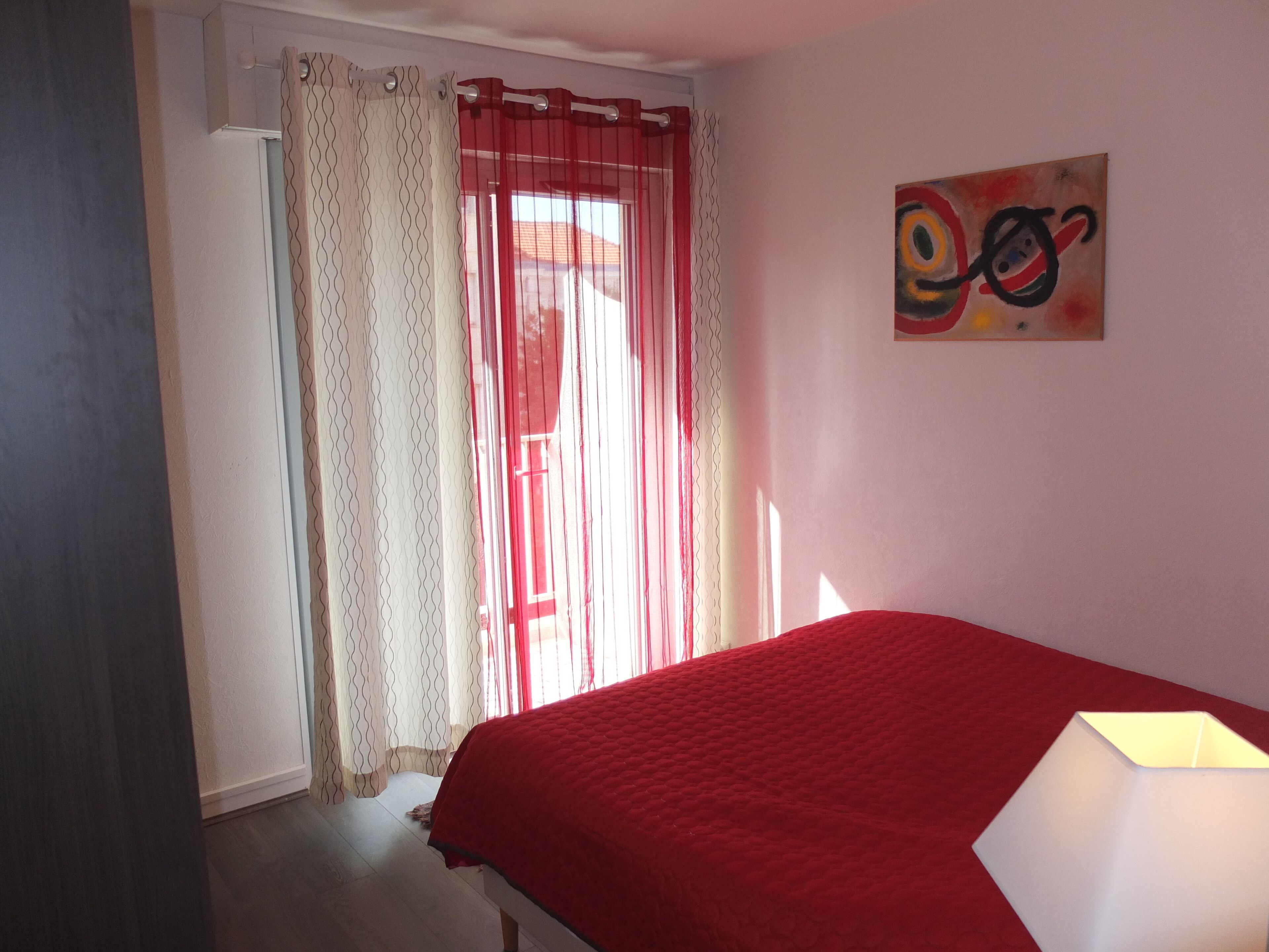 Une chambre à coucher avec un lit, une fenêtre ornée de rideaux rouges, un tableau accroché au mur et une lampe sur la table de chevet.