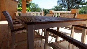 A wooden table and chairs under a pergola, with a view of trees and a white fence in the background.
