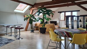 Een eetgedeelte met een hoge plafond, houten balken, een grote plant, gele stoelen en een tafel met een geruit tafelkleed.