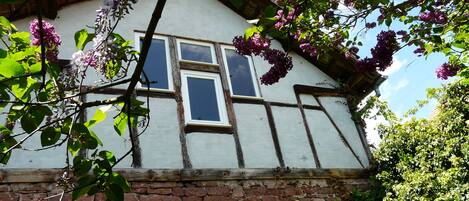 Una casa tradicional de entramado de madera con base de piedra, dos ventanas y árboles en flor al frente.