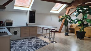 Een compacte keuken met een wasmachine, een magnetron en een inbouwoven. Twee dakramen, een grote potplant en een bank in een woongedeelte.