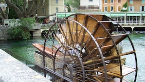 A large water wheel structure near a river, with buildings in the background. - cccccccccc (Fontaine-De-Vaucluse)