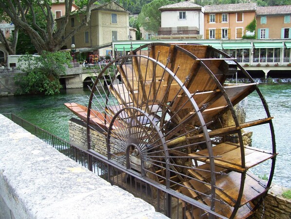 A large water wheel structure near a river, with buildings in the background. - cccccccccc (Fontaine-De-Vaucluse)