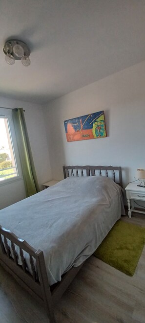 A bedroom with a bed, a bedside table, a lamp, a window with curtains, and a wall painting.
