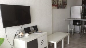 Ett modernt vardagsrum med en platt-TV som är monterad på väggen, ett vitt skåp med förvaringsutrymme, en vit bänk och ett litet bord med en stol.