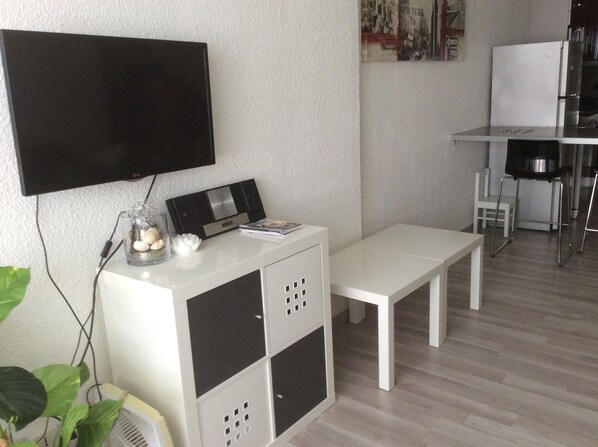 Een moderne woonkamer met een flatscreen televisie aan de muur, een wit kastje met opbergruimte, een witte bank en een kleine tafel met een stoel.