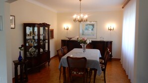 Ein Esszimmer mit Tisch für vier Personen, umgeben von Holzsesseln. Eine Chandelieber hängt von der Decke, und links befindet sich ein Schrank mit Glastüren.
