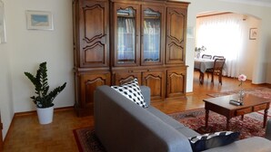 Ein Wohnzimmer mit grauem Sofa, einem Holzschrank, einem Esstisch mit Stühlen und einer Topfpflanze.