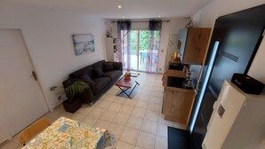 A compact living space with a sofa, a small table, a kitchen area, and a sliding glass door leading to a balcony. - Little house with pool 4 people (Payzac)