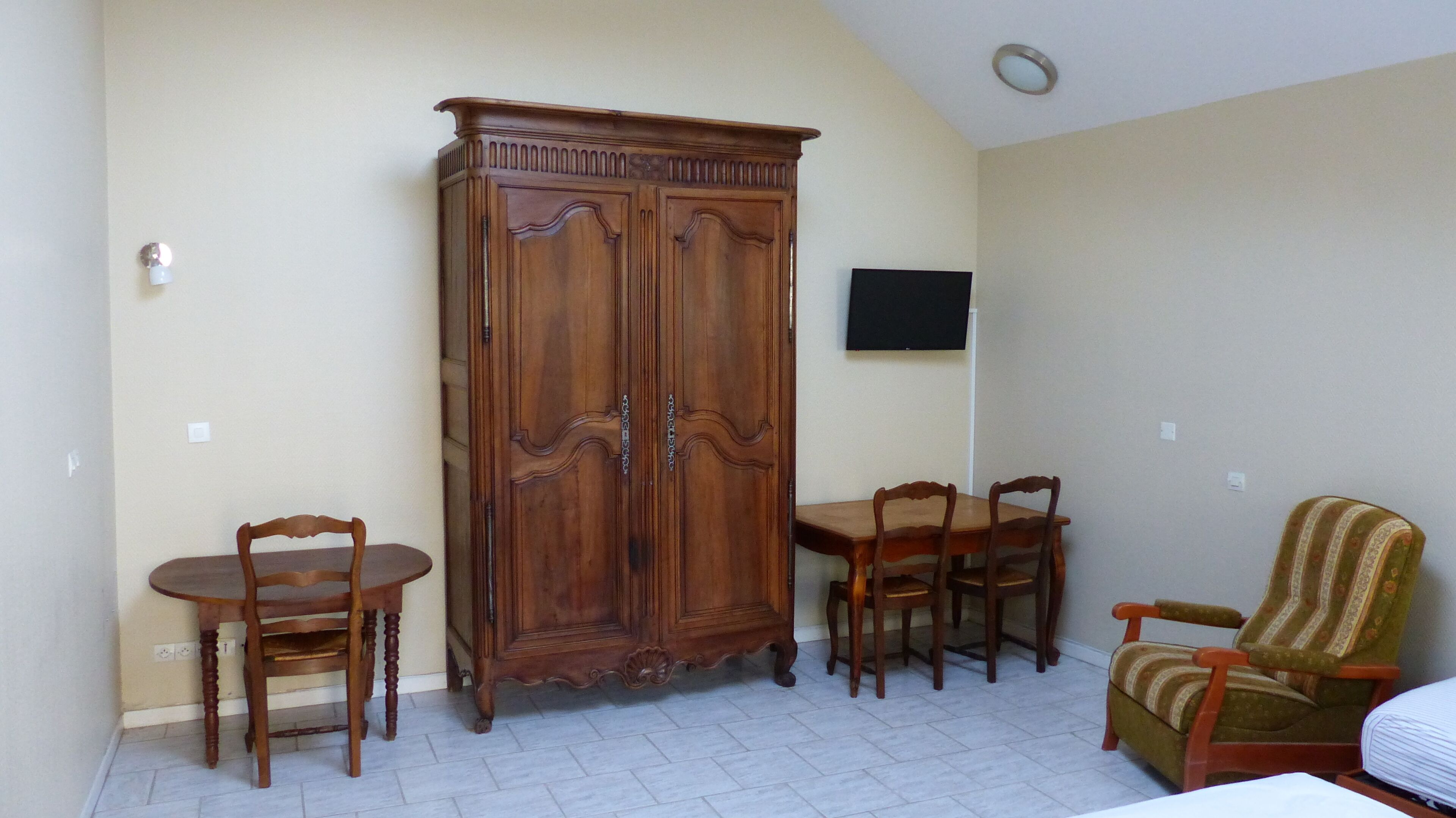 Une pièce comprenant un meuble en bois, une table à manger avec des chaises, un téléviseur fixé au mur et un fauteuil à motifs.
