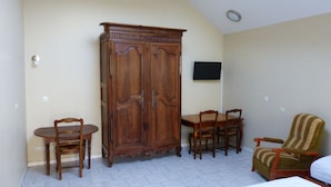 Una habitación con un armario de madera, una mesa de comedor con sillas, un televisor montado en la pared y un sillón con estampado.