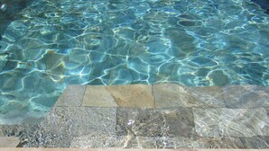 Uma piscina retangular com água cristalina, ladeada por uma borda de pedra e uma área gramada.