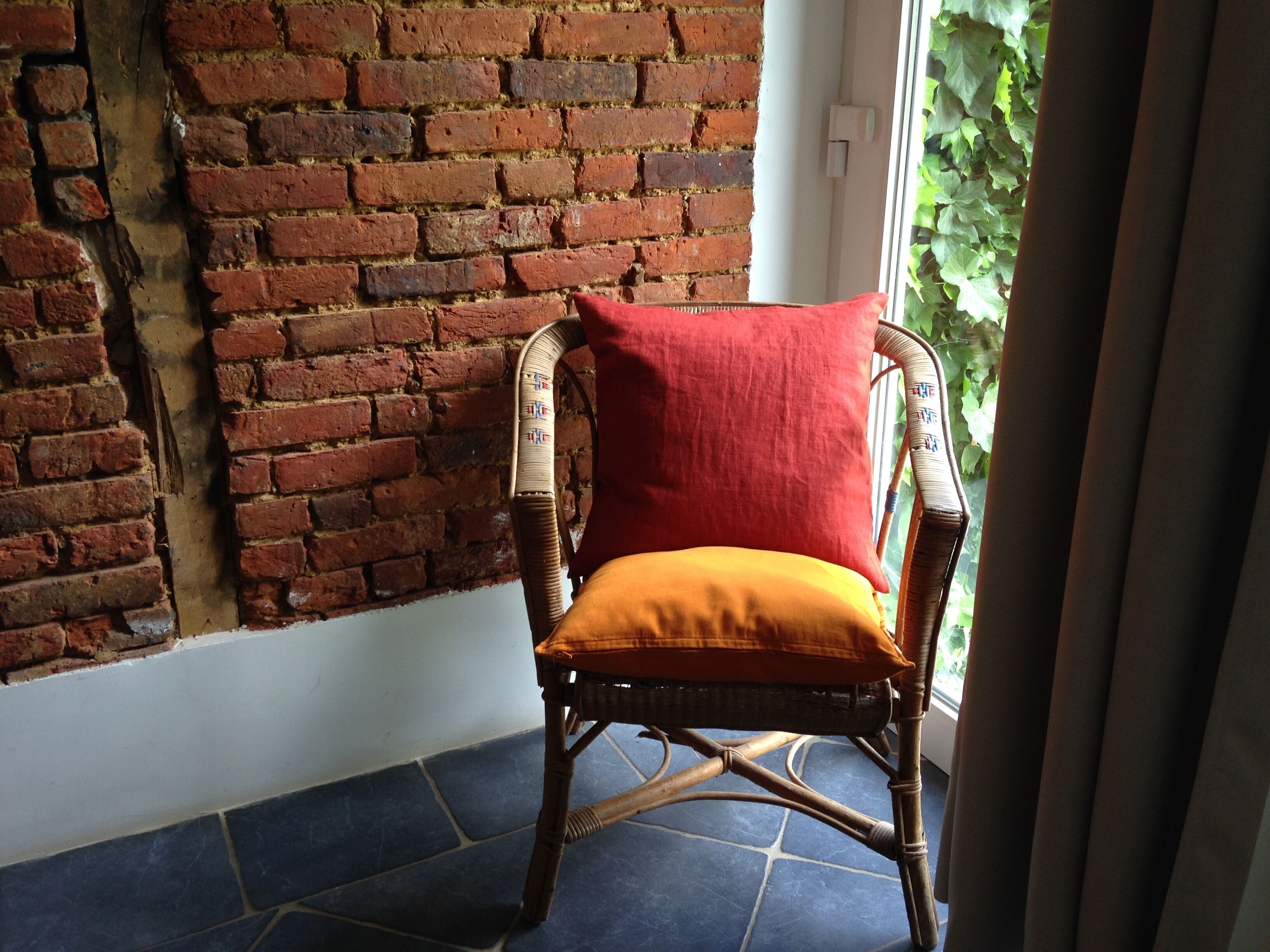 A wicker chair with a red cushion and an orange cushion against a brick wall.