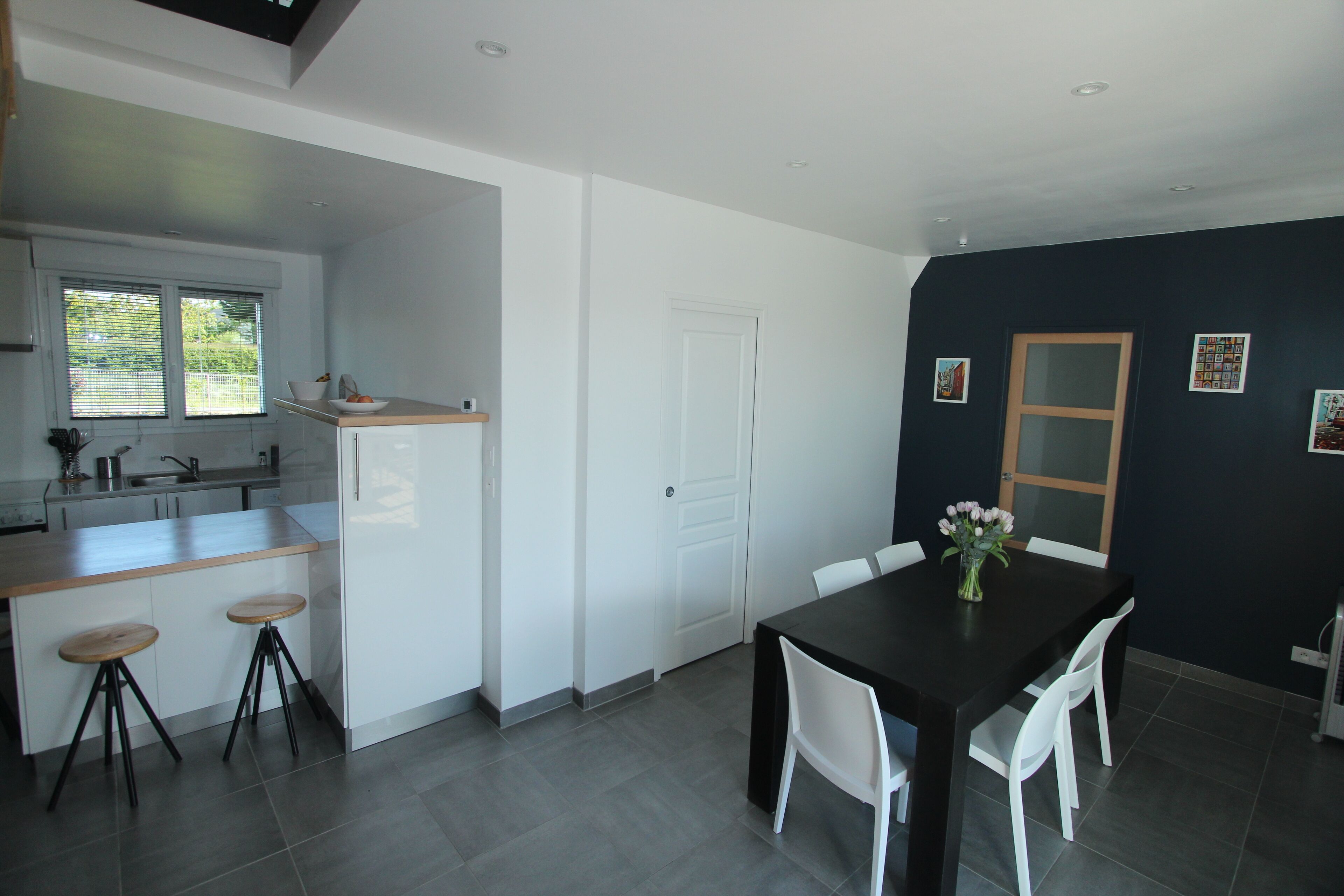 A modern kitchen with a dining area, featuring a large island with stools, a dark dining table with chairs, and a door leading to another room.