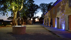 An outdoor pathway leading to a building with a rustic facade, a large tree with a metal structure in the foreground, and a sunset in the background. - Le 7 Mas - Suite Angélique & Adrienne (Saint-Andiol)