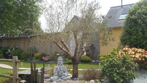 Ein Garten mit Buddha-Statue, Holzzaun, Baum und Haus im Hintergrund.