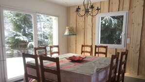 Un comedor con una mesa dispuesta para seis personas, sillas de madera, una araña y un ventanal con vistas a árboles cubiertos de nieve.