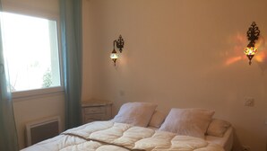 Una cama con colcha, dos lámparas de pared, una mesita de noche y una ventana con cortinas.
