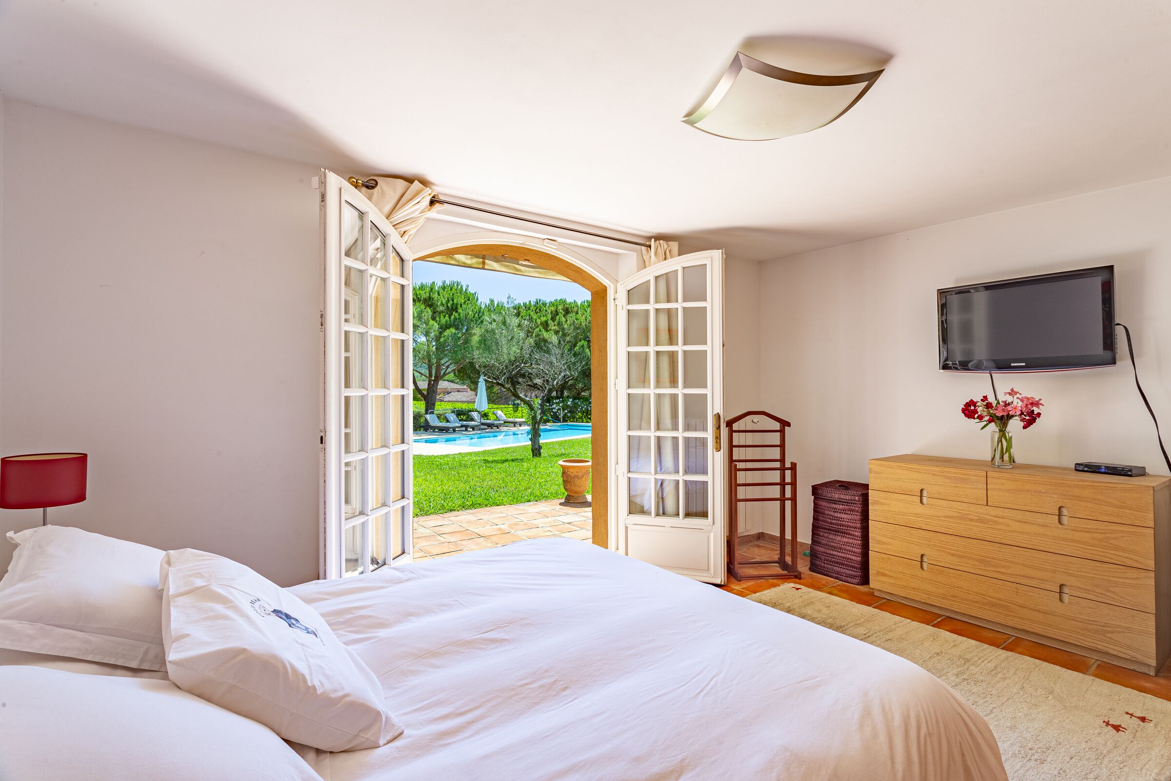 Quarto com cama, cômoda, televisão e porta aberta para área da piscina.