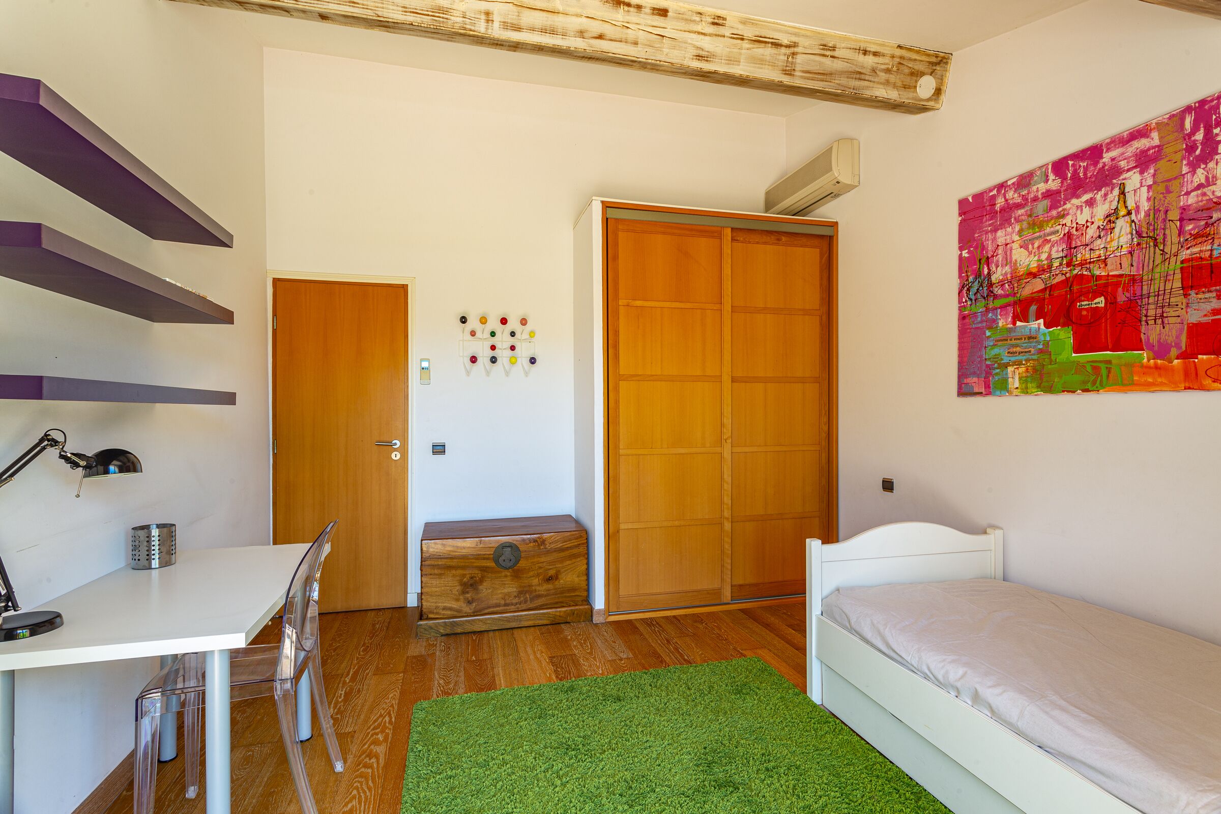 A bedroom with a bed, a desk with a chair, a wooden wardrobe, and a colorful abstract painting on the wall.
