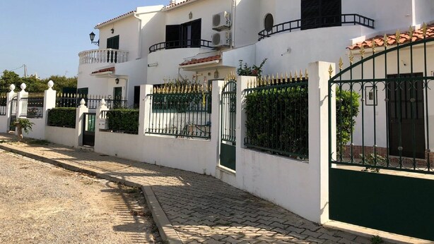 A row of white buildings with balconies, a gated entrance, and a paved street.