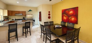 A modern kitchen with a dining area, featuring a large island with bar stools, a stainless steel refrigerator, and a painting of tomatoes on the wall.