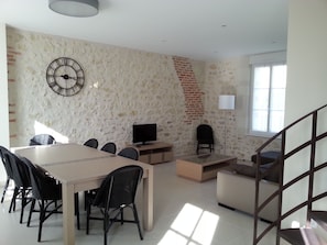 A modern living room with a stone wall, a large clock, a TV, a dining table with chairs, a sofa, and a staircase. - Gîte du Domaine du Haut Perron near the Beauval ZooParc (Thésée)