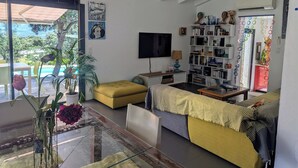 A modern living room with a glass coffee table, a yellow ottoman, a striped sofa, and a bookshelf filled with books and decorative items. - Villa in Lumio, large swimming pool, wooded park   (Lumio)