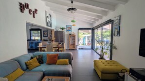 A spacious living room with a sofa, ottoman, and a coffee table. There is a kitchen area with cabinets and a dining table. The room has a large window and a ceiling fan. - Villa in Lumio, large swimming pool, wooded park   (Lumio)