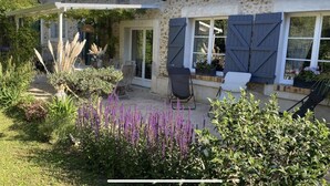 A stone house with a covered patio, outdoor seating, and a garden with purple flowers.