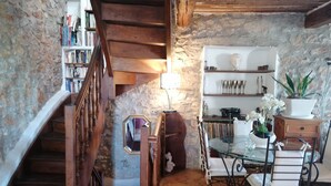 A rustic interior with a wooden staircase, stone walls, a glass table with white chairs, and a bookshelf with books and decorative items.