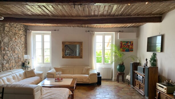 A spacious living room with a stone wall, a wooden ceiling, and a hexagonal tiled floor. There is a beige sofa, a coffee table, and a television on a wooden stand.