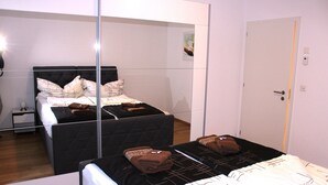 A bedroom with a mirrored wardrobe, a bed with a black quilted headboard, and a bedside table with a lamp. - Modern, new 3.5-room apartment in brig with fantastic views of the mountains (Brig-Glis)
