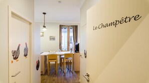 Un couloir étroit mène à une salle à manger où se trouvent une table, des chaises et un luminaire suspendu.